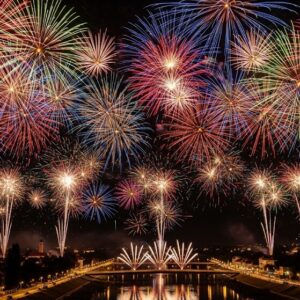 夜空全体に広がる、赤、金、青、緑の色鮮やかな花火。川面に街の灯りが反射している。